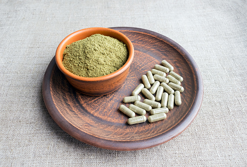 Supplement kratom green capsules and powder on brown plate. Herbal product alt-medicine kratom is  opioid. Home alternative pain remedy, opioid addiction, dangerous painkiller, overdose. Close up