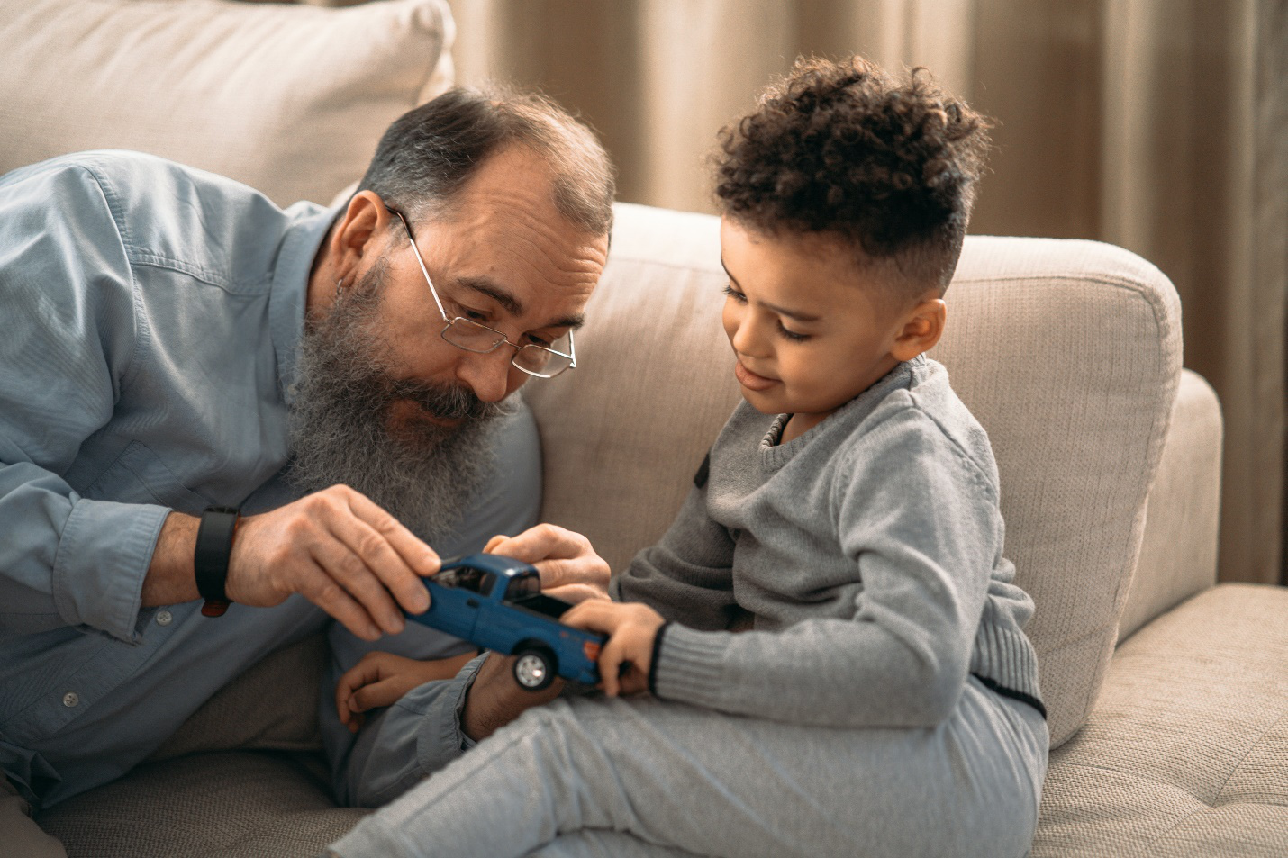 A bearded grandfather in blue sits with his grandson on a sofa, examining a blue pickup truck