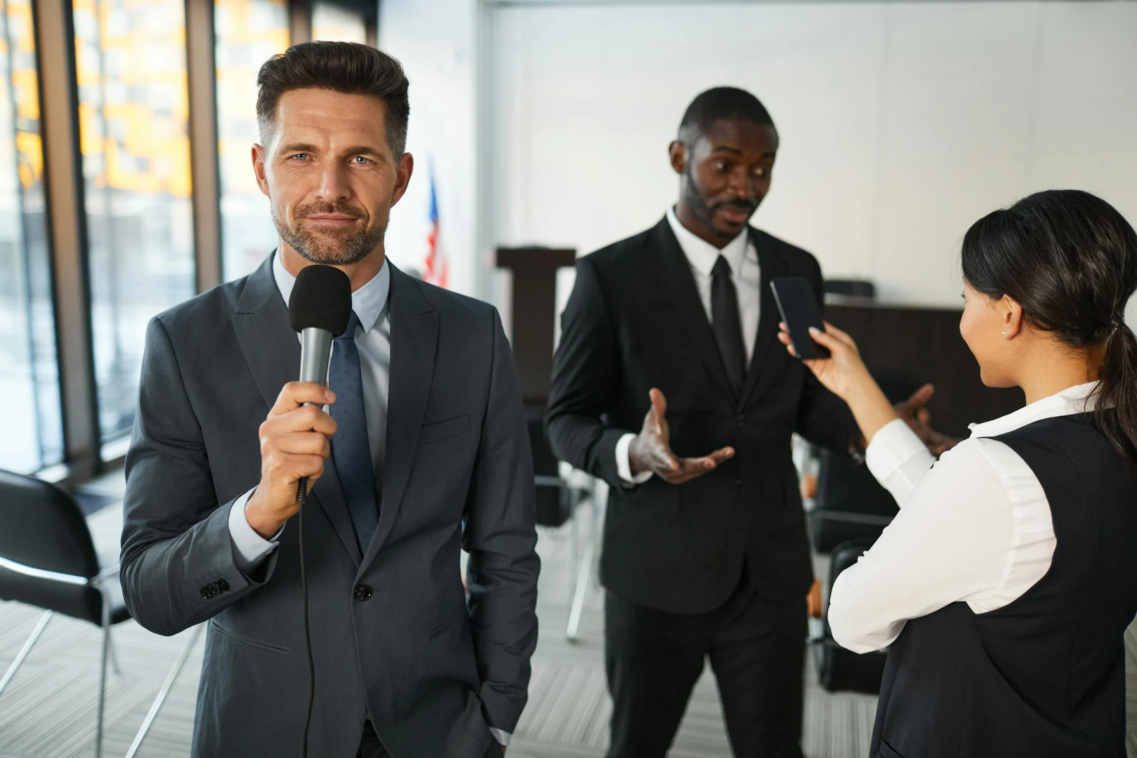 A broadcast journalist, wearing a suit and holding a microphone, stands in a studio.