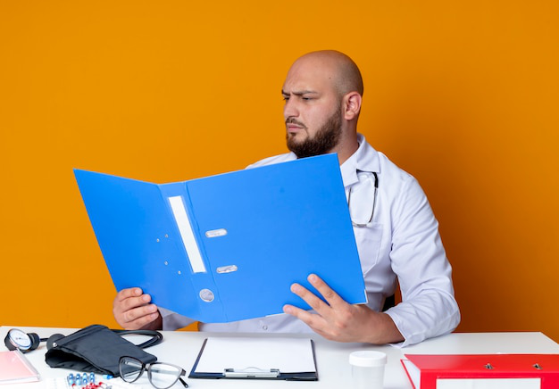 A doctor in a medical robe and stethoscope reviewing a medical file on a desk.